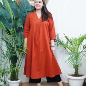 Model wearing a solid rust-colored A-line kurta with V-neck embroidery and side pockets, paired with black pants.