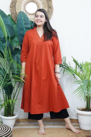 Model wearing a solid rust-colored A-line kurta with V-neck embroidery and side pockets, paired with black pants.